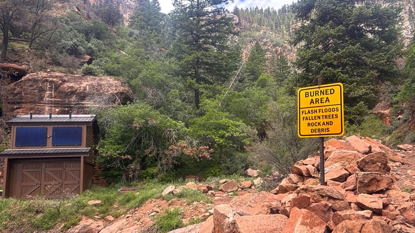 Composting toilet by a trail in forested area with sign warning hikers of potential hazards.