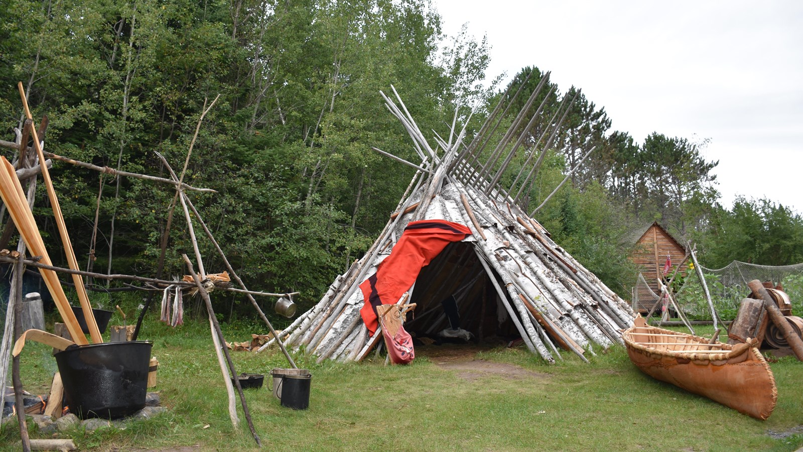 Birch bark tipi, canoe, and other Ojibwe cultural items on a grassy area.