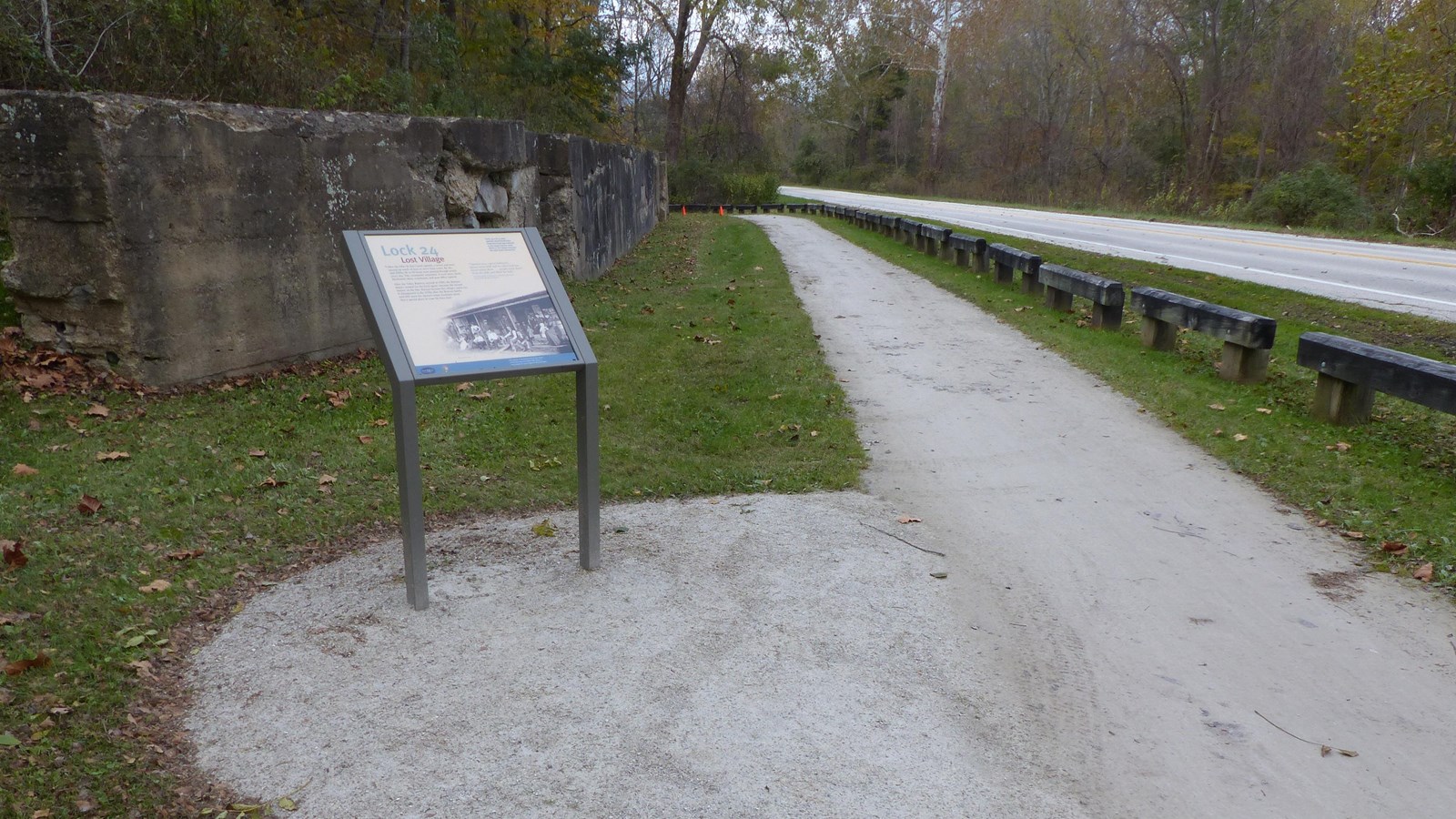 Exhibit panel by a stone wall faced with crumbling cement near an unpaved trail.