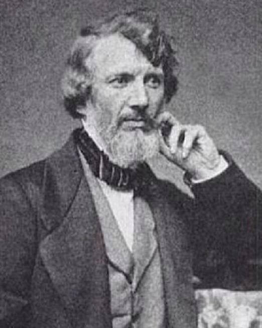 Man in suit sits with elbow resting on table. Hair combs to 1 side & has trimmed beard & mustache.