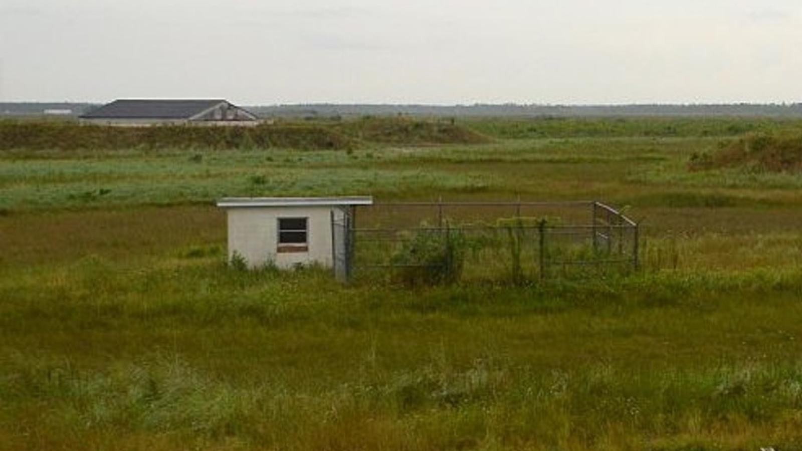 A small white building with adjacent squared in fence. Flat landscape is covered with grasses.