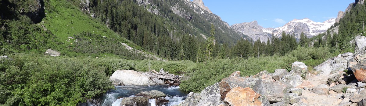 A river runs beside a rocky trail in the middle of a mountain canyon.