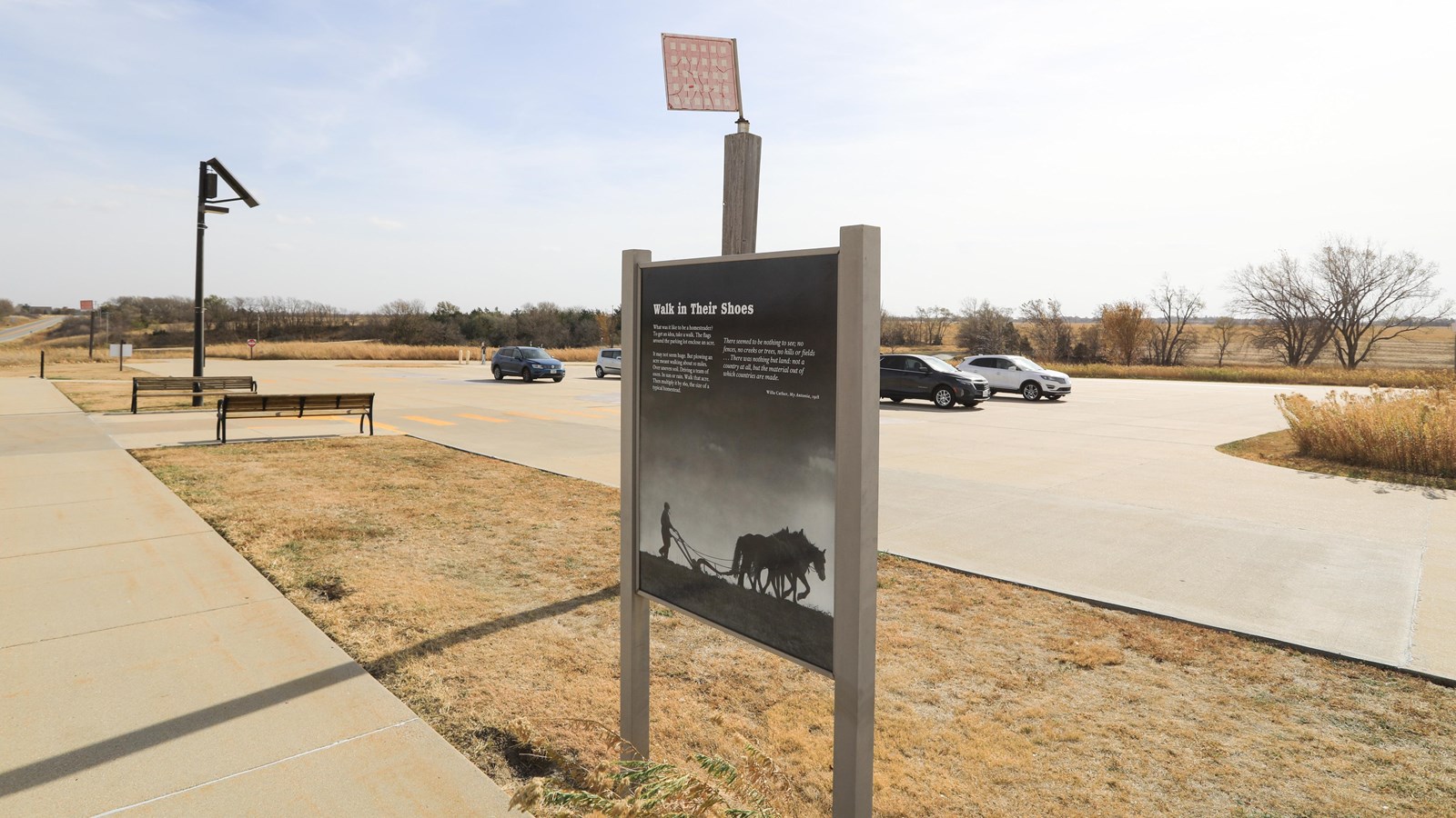 Informational sign stands in front of parking lot. Lot is surrounded by checked flags.