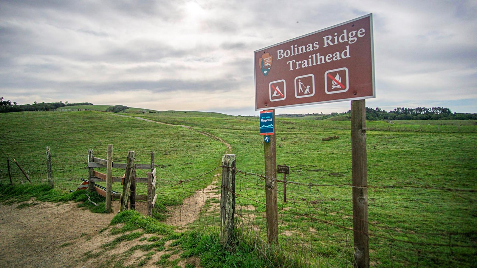 A trailhead sign: Bolinas Ridge Trailhead. The trail begins beyond a wooden cattle gate .