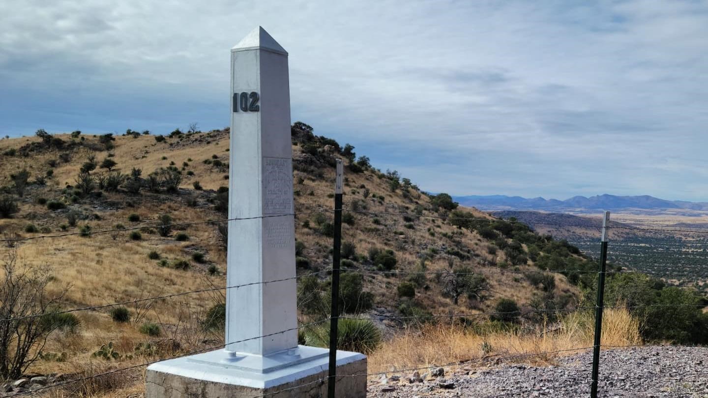 A silver obelisk sits on a concreate base behind a barbed wire fence.