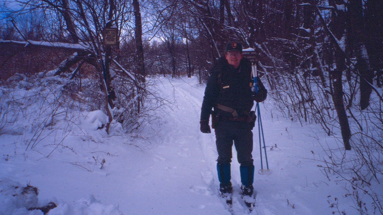 A park ranger in green uniform, skiing along a snowy forest trail.