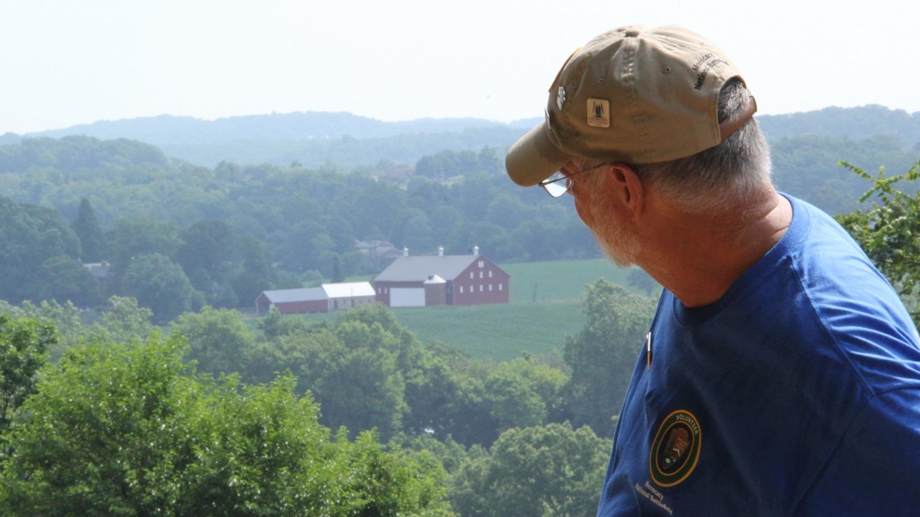 A white male faces away from camera and looks at a red barn and farm buildings in distance below.