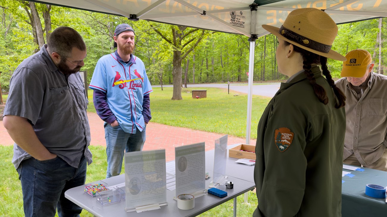 A female park ranger speaks to three men under a tent with a table set up.