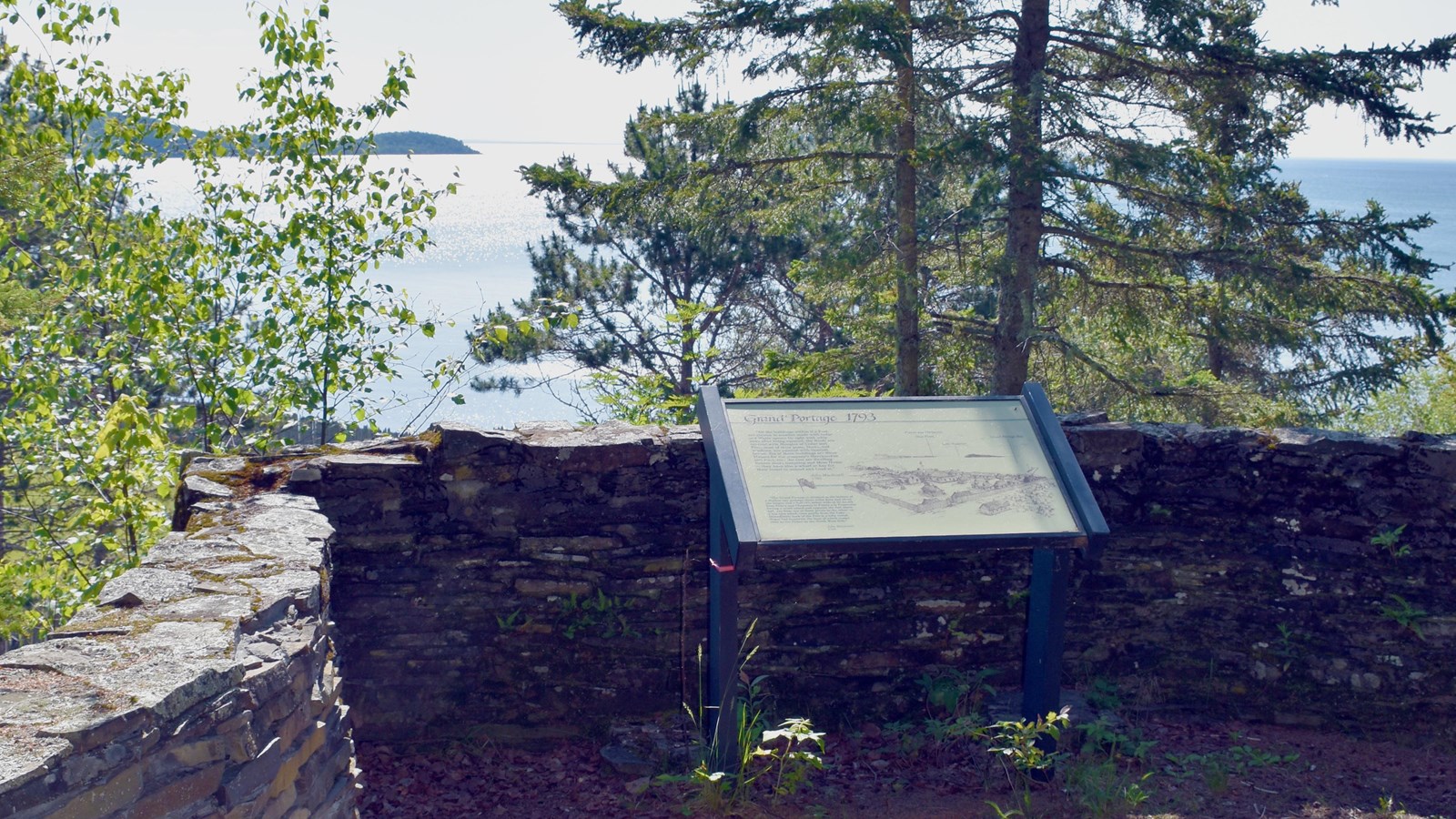 Interpretive wayside in front of a low stone wall with a view of a bay beyond.