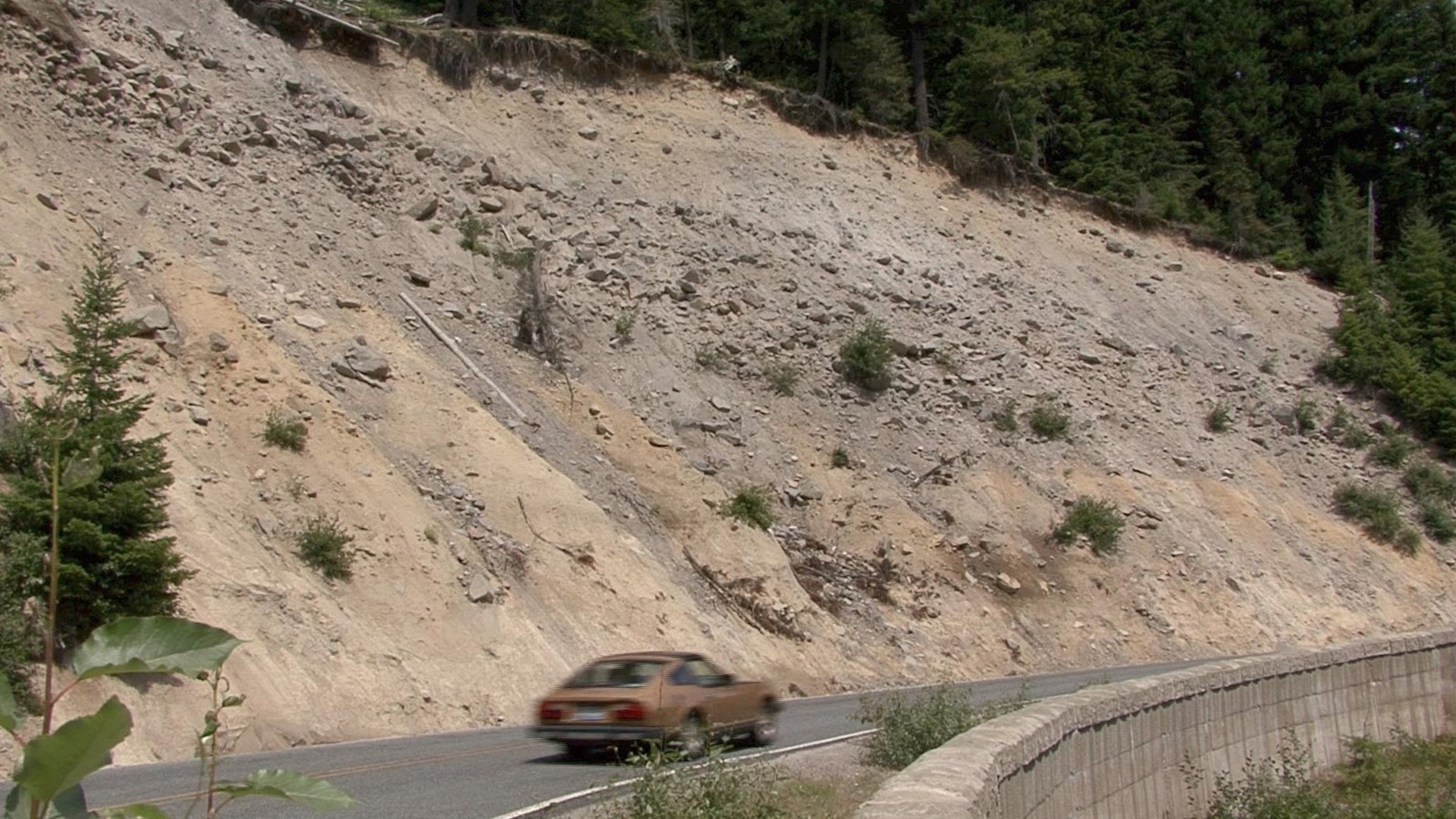 A car drives along a curved road next to an exposed hillside road cut showing layers of sediment.