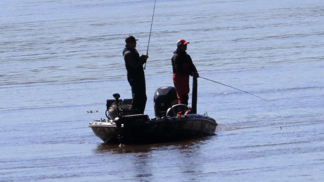 Two men with fishing poles standing on a small boat in the water,