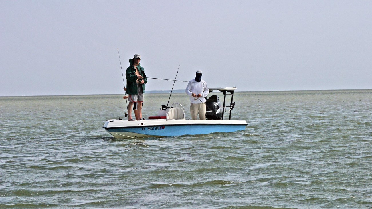 Two anglers stand on a light blue fishing boat in gentle waters. They hold fishing poles in hands
