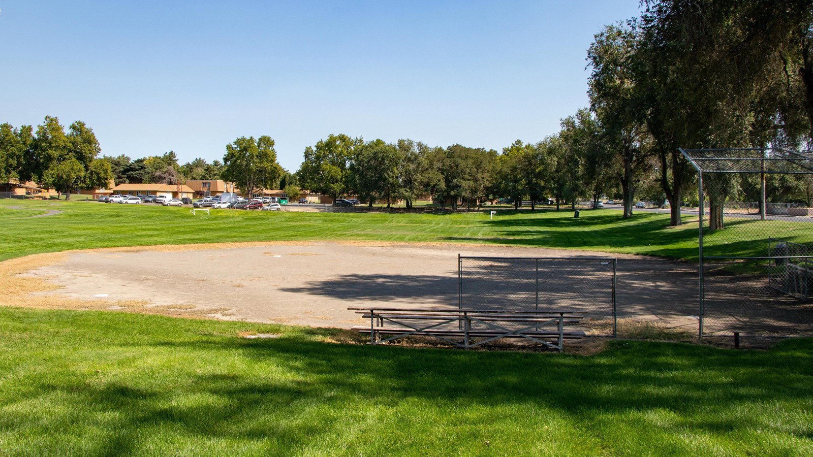 A large sandy area with metal benches surrounded by mowed, grassy green area lined with trees.