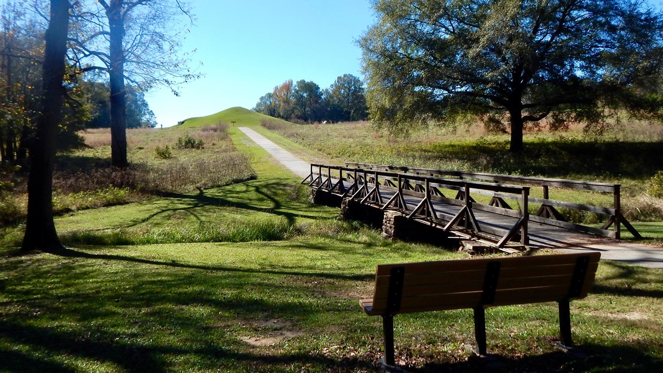 A park bench overlooking the paved main trail leading towards the Earth Lodge.