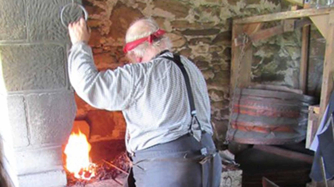 Man in 19th century clothes working over a blacksmith\'s forge.