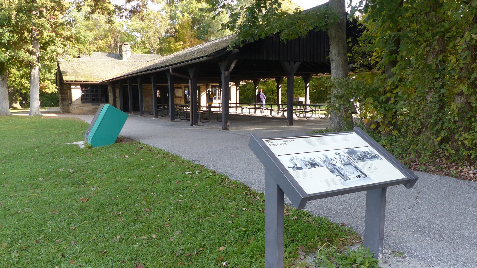 Graphic panel along a paved path to a rustic shelter; rows of picnic tables under its roofed porch.