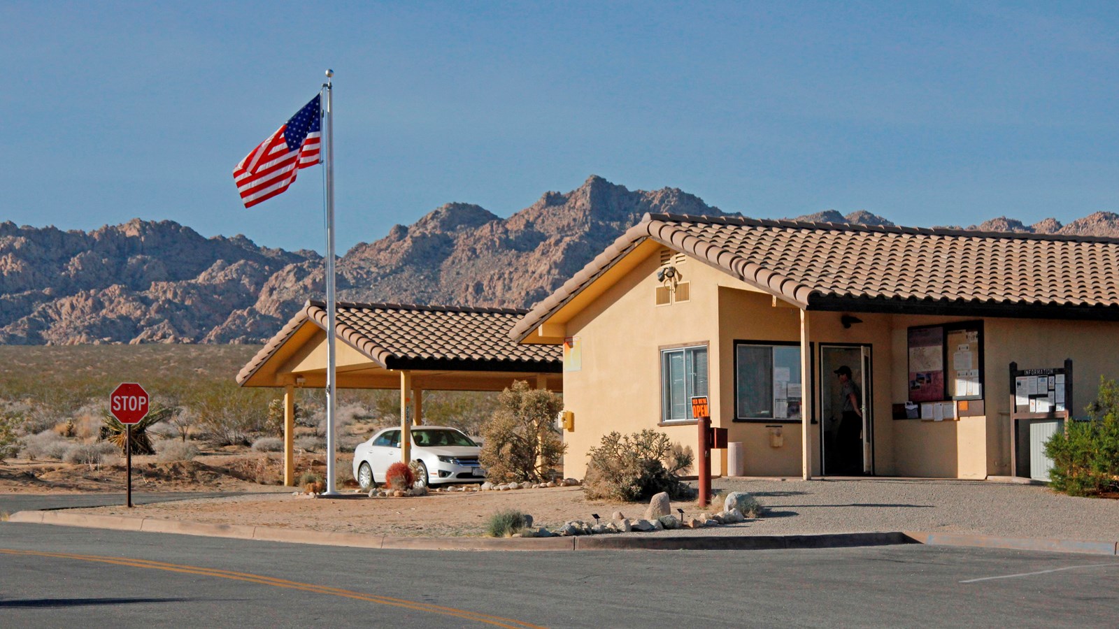 A tan one story building with stucco roof by a parking lot with rocky mountains in the background.