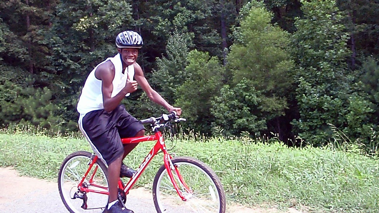 Cyclist giving a thumbs up as he rides on trail with woods in the background.