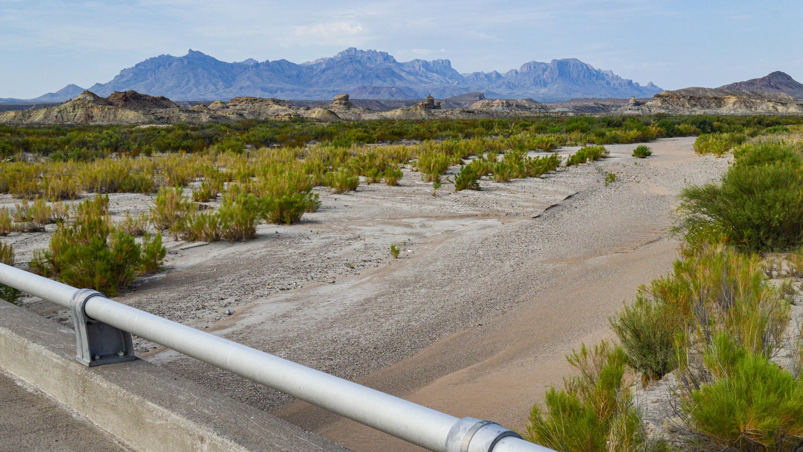 A concrete bridge spans a wide drainage, partially scoured of vegetation by floods.