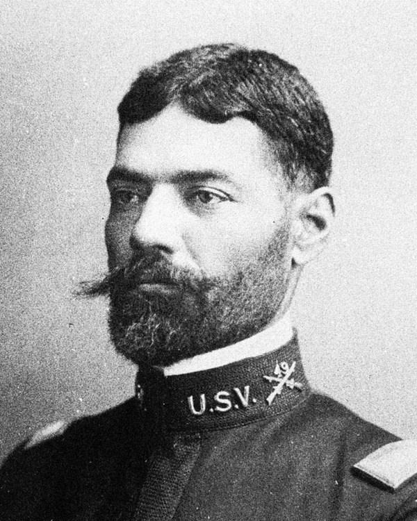 Black and white photograph of African Man from the chest up. He has on military uniform from 1890s