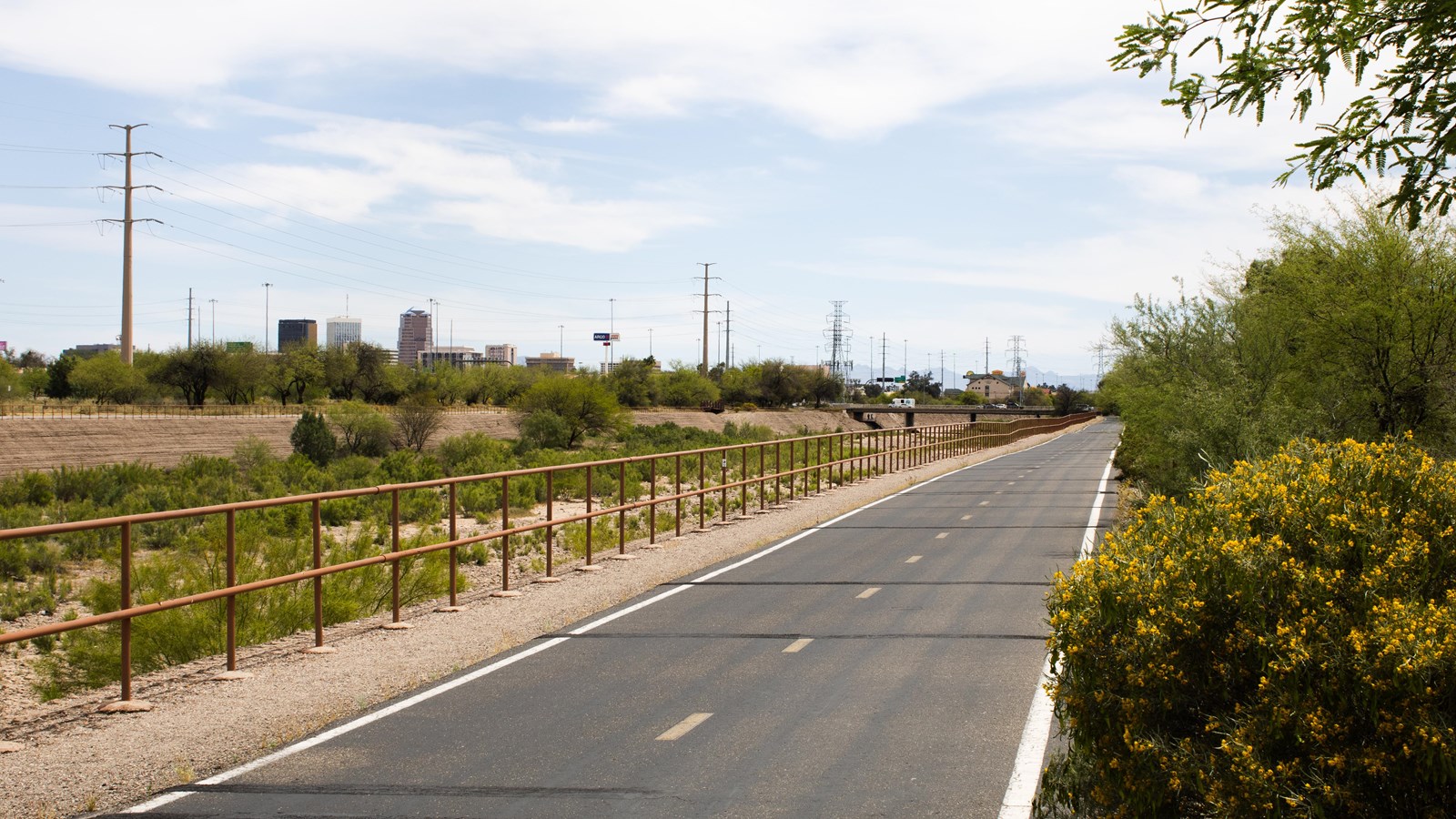 A paved bike path has green brush on the right and a red metal fence in front of a dry riverbed