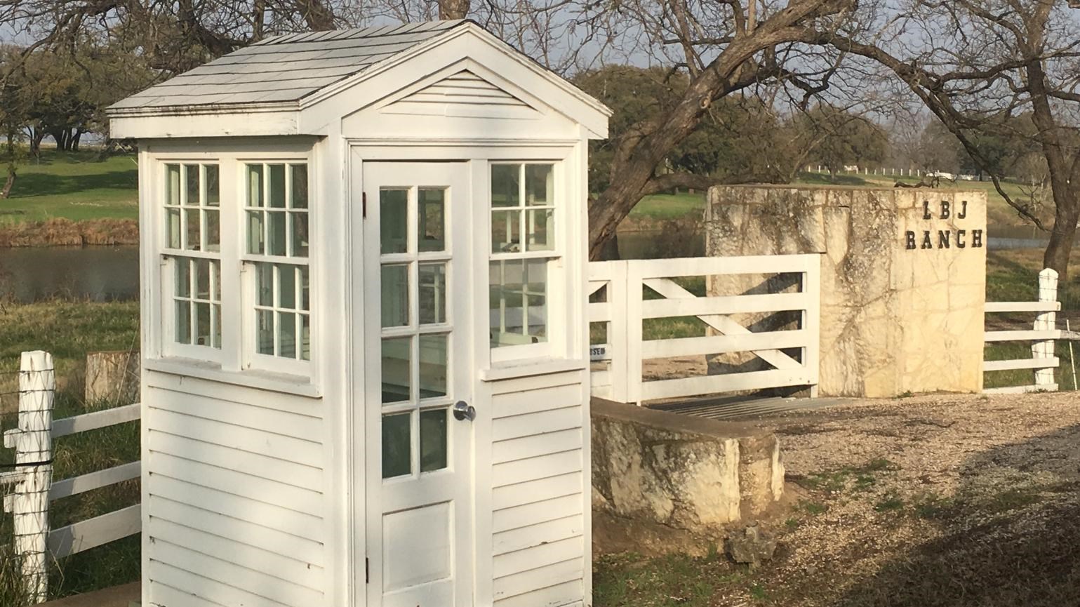 A white guard shack stands in front of fenced property near a stone wall that displays 