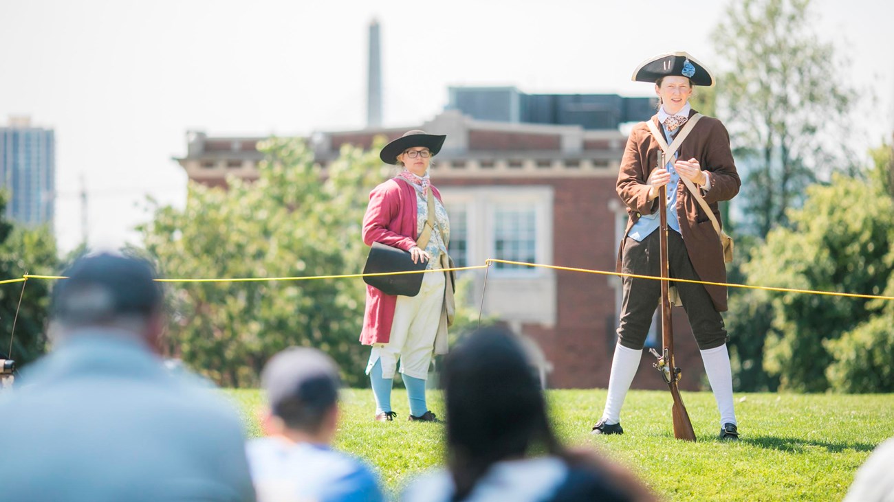 Two Park rangers dressed in period clothing, one has a musket to her side and speaks to the audience