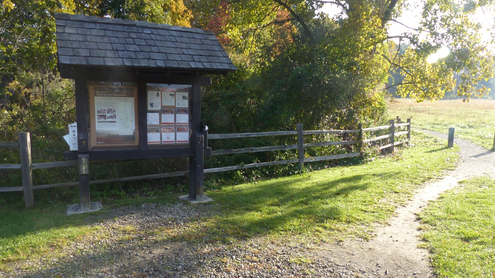 Trailhead bulletin board, left, stands along a rail fence. Unpaved path heads away beside a field.
