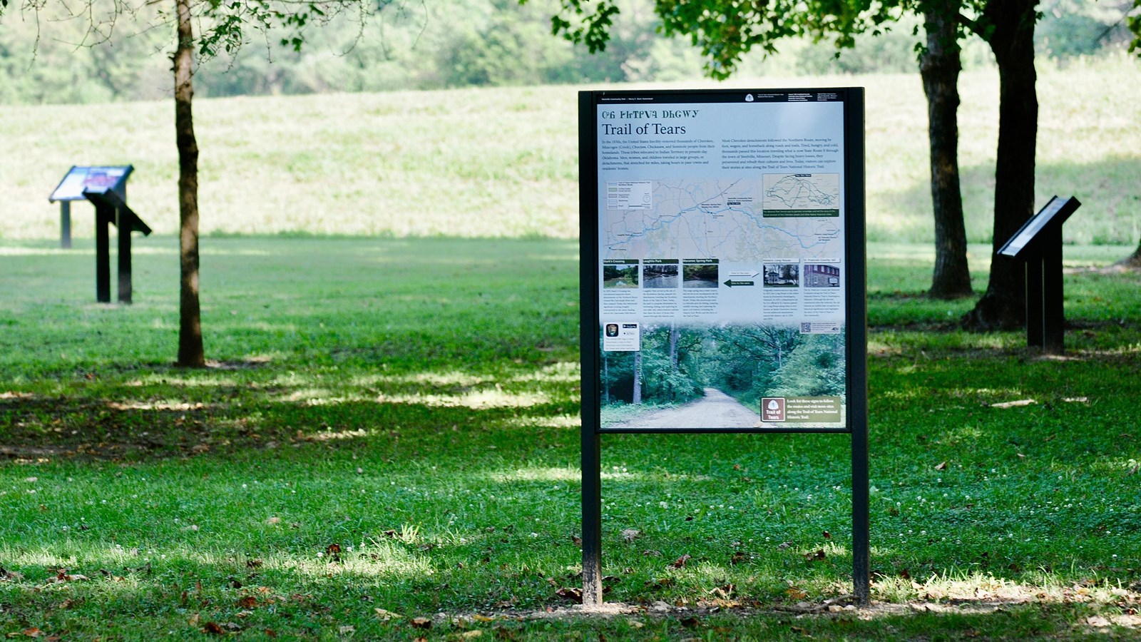 An upright wayside exhibit about the Trail of Tears is in front of a green, grass field.