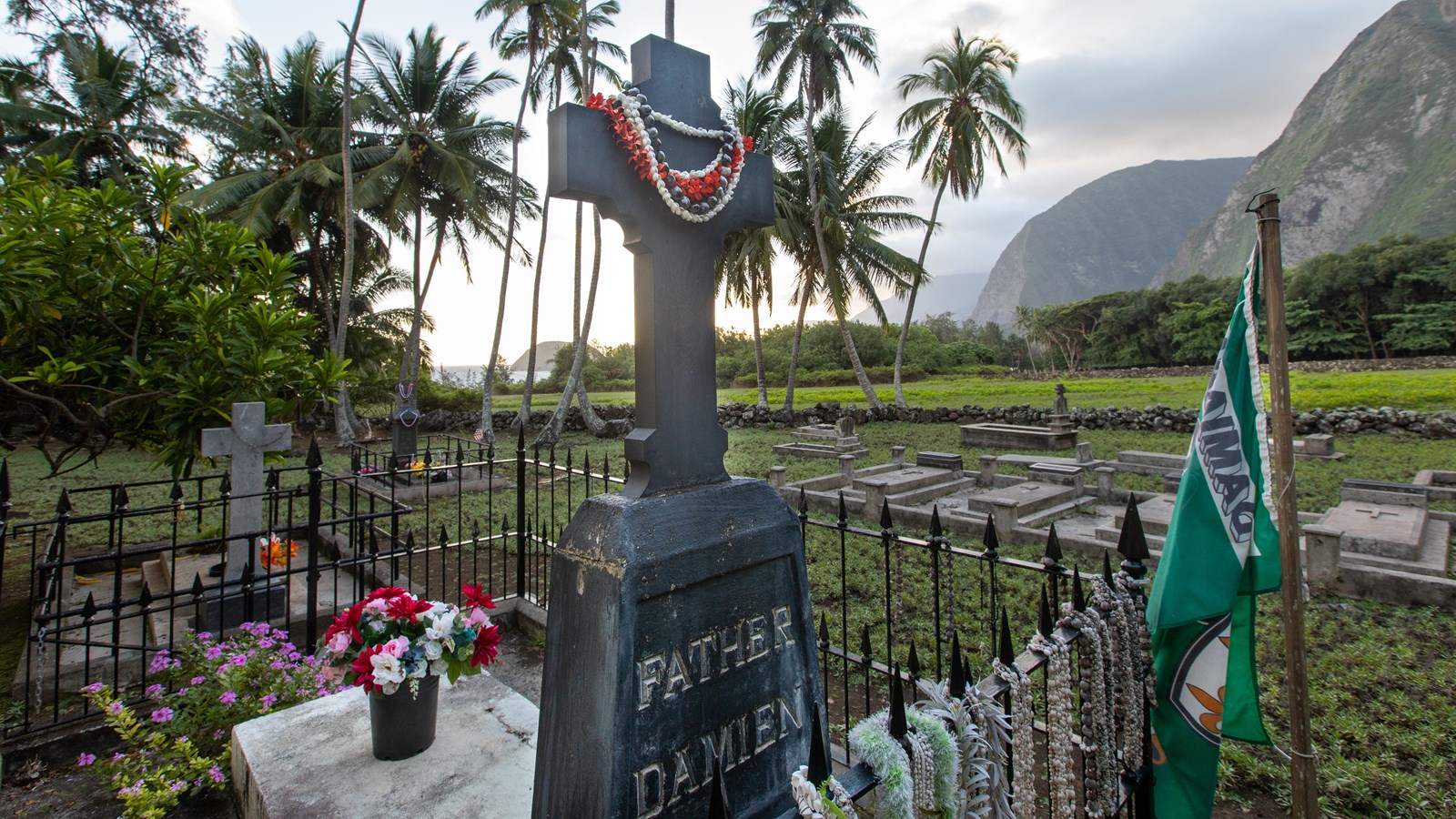 A large wooden cross on a grave marker that reads Father Damien decorated with leis. 