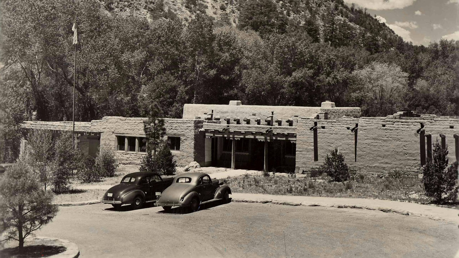 a stone building with cliffs behind and 2 old cars parked in front