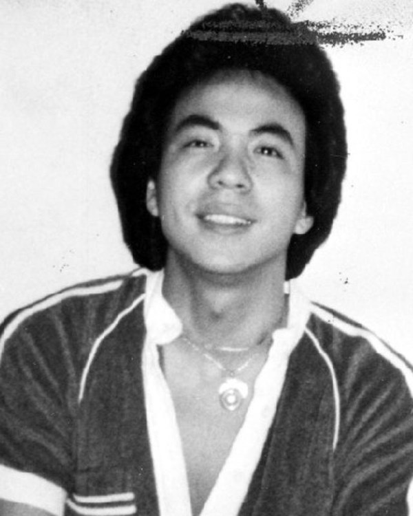 Black and white image of Asian man smiling at the camera. They are wearing a dark V-neck shirt.