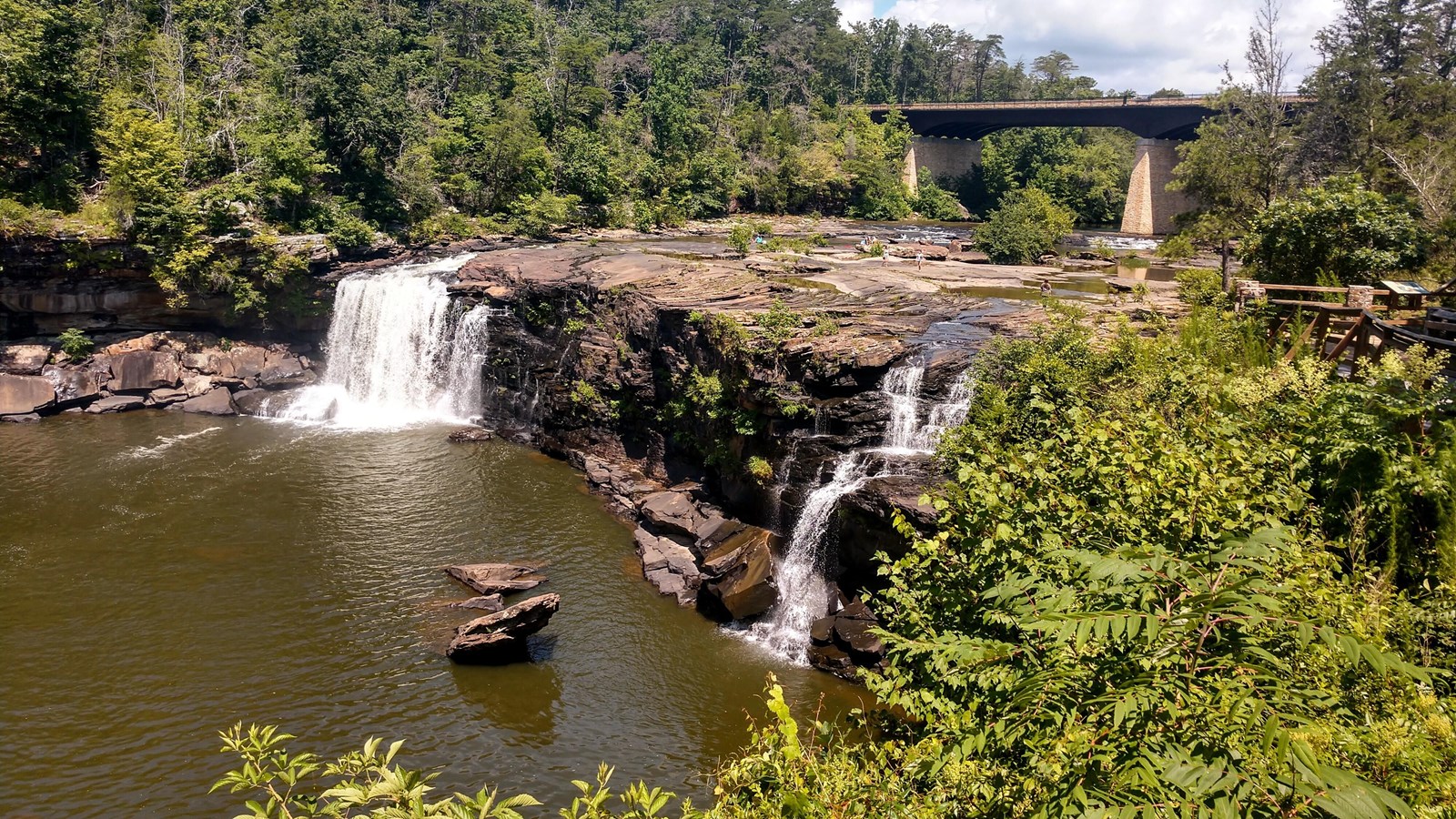 A large, multi pronged waterfall tumbles off a ciffside into a pool of water, surrounded by forest.