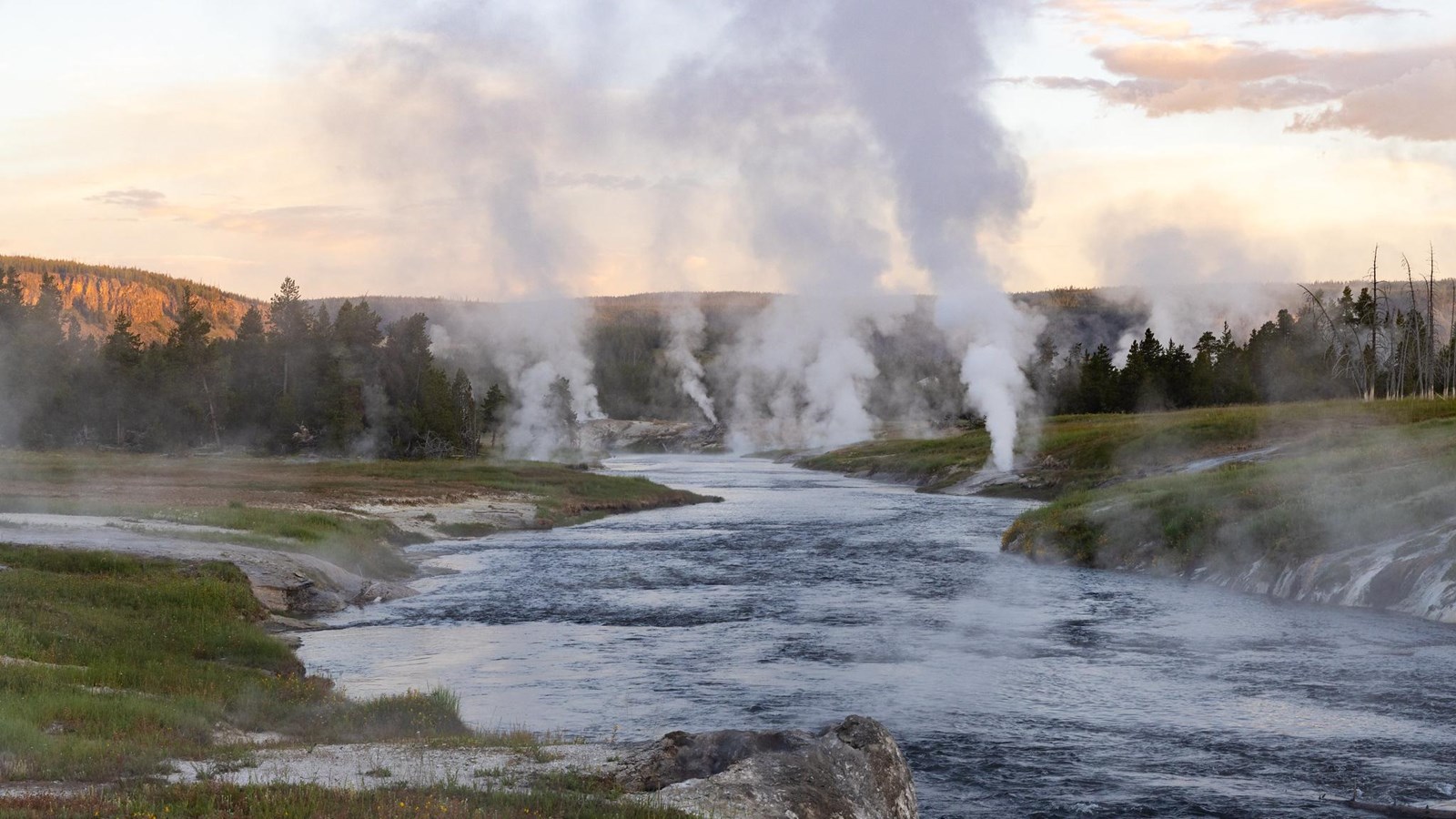 Steam rises from thermal features along the banks of a river at sunrise.