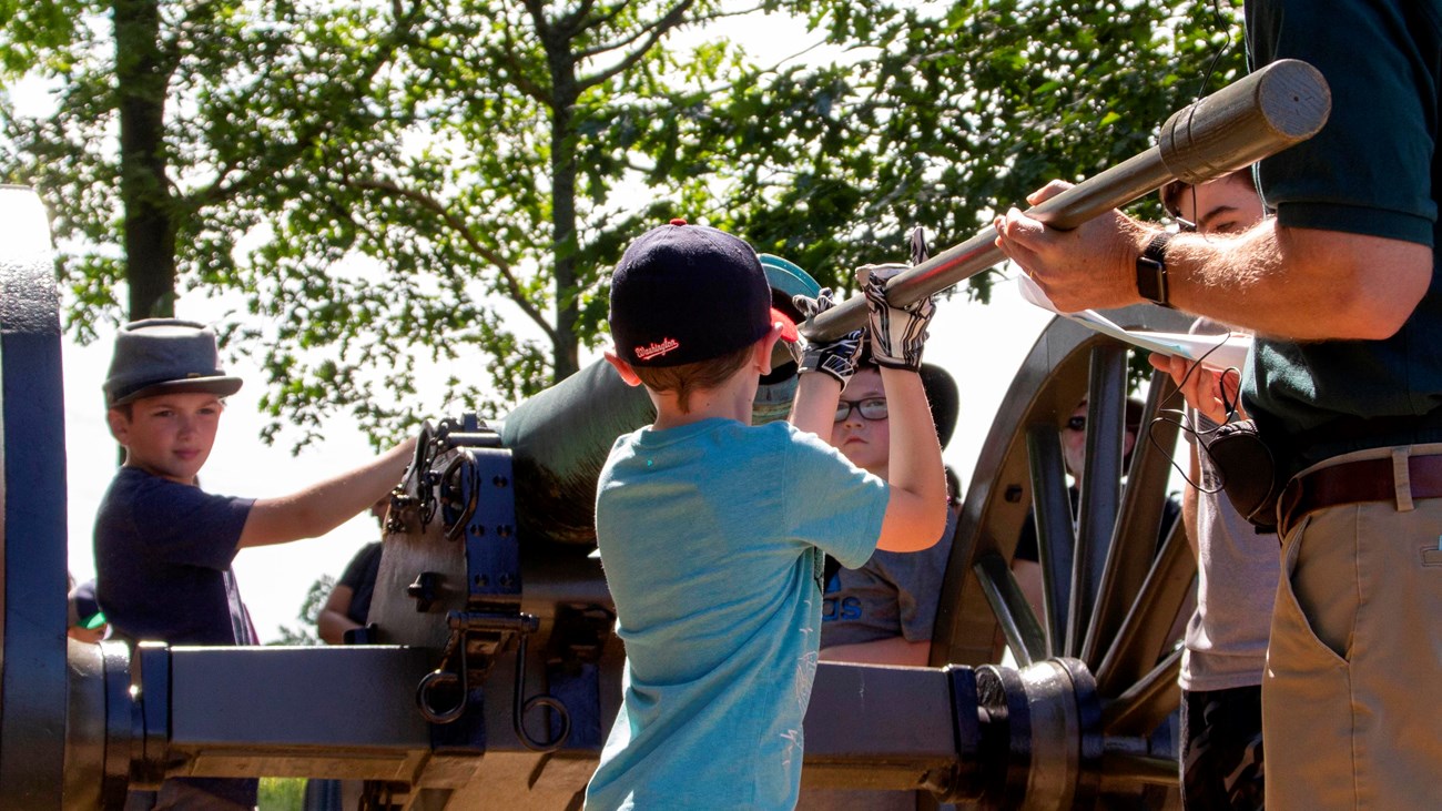 A child stands near a cannon near a park volunteer who instructs him on its use.