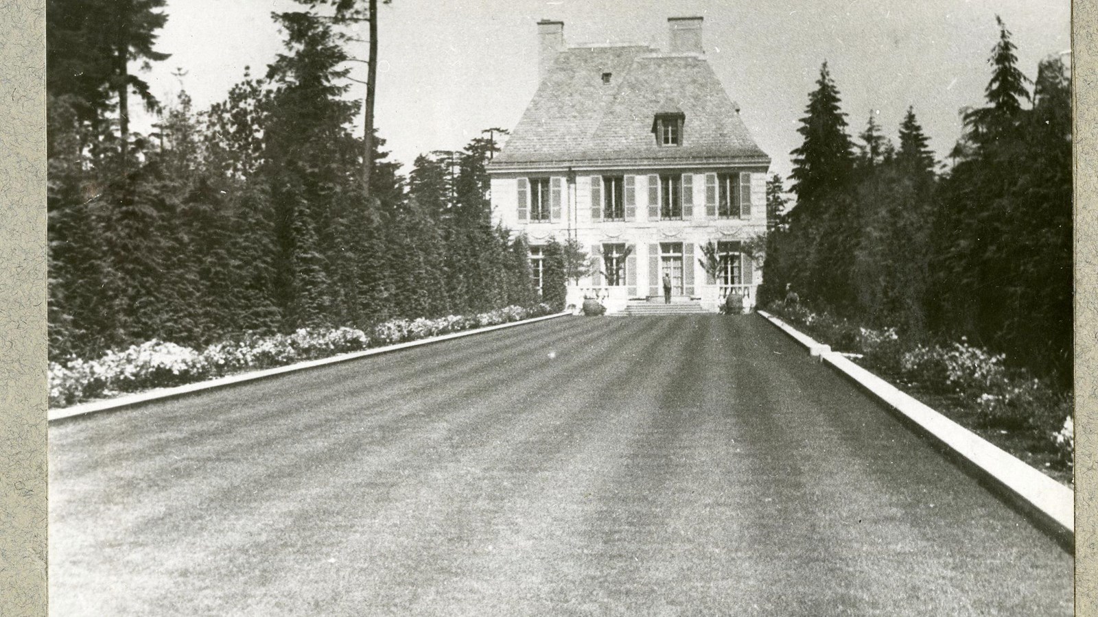 Black and white of flat grassy area with large trees on side, large house at end