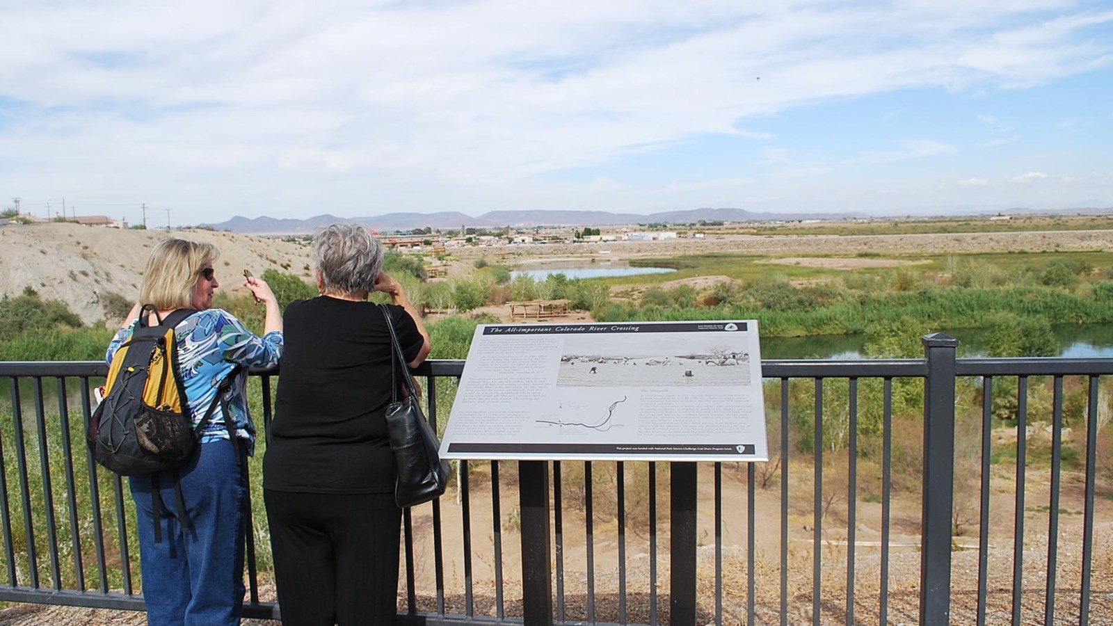 Two women stand against a black metal fence overlooking a river in a desert