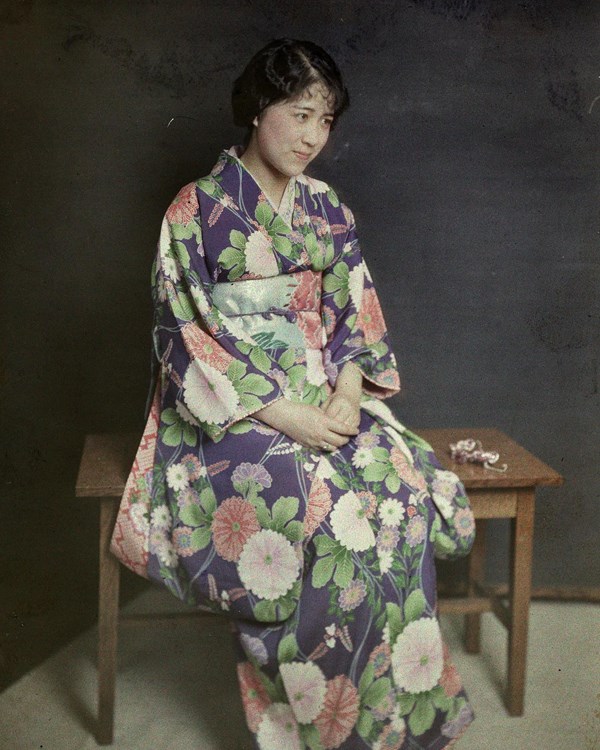 Japanese woman wearing navy kimono with pink and white flowers and green leaves sits on wooden stool