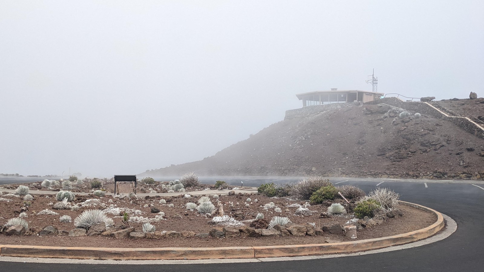 a blacktop with a center island of cinder and small plants a hill and building in distance in fog
