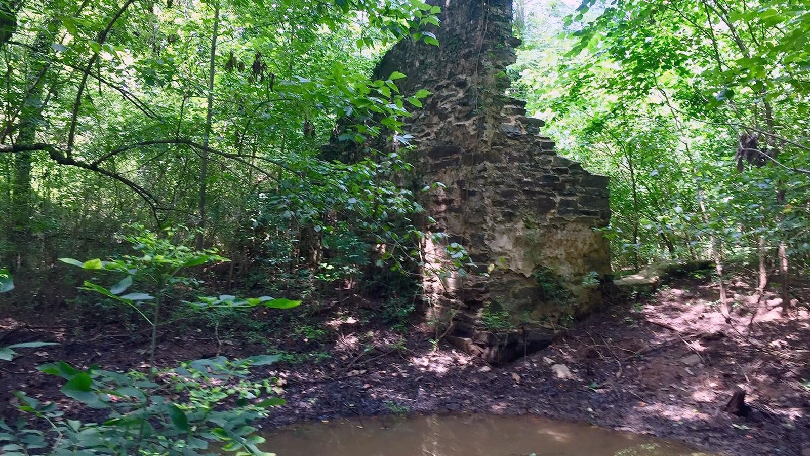 Corner of stone walls of mill with pool of water in the foreground and surrounded by woods.