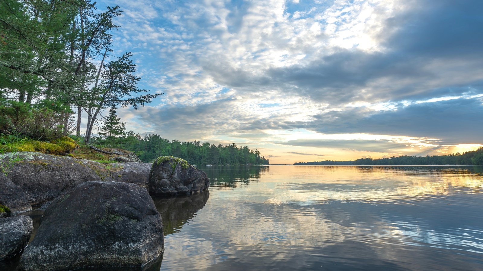 A rocky shoreline with trees on the left with a lake on the right side. Wispy clouds in the sky.