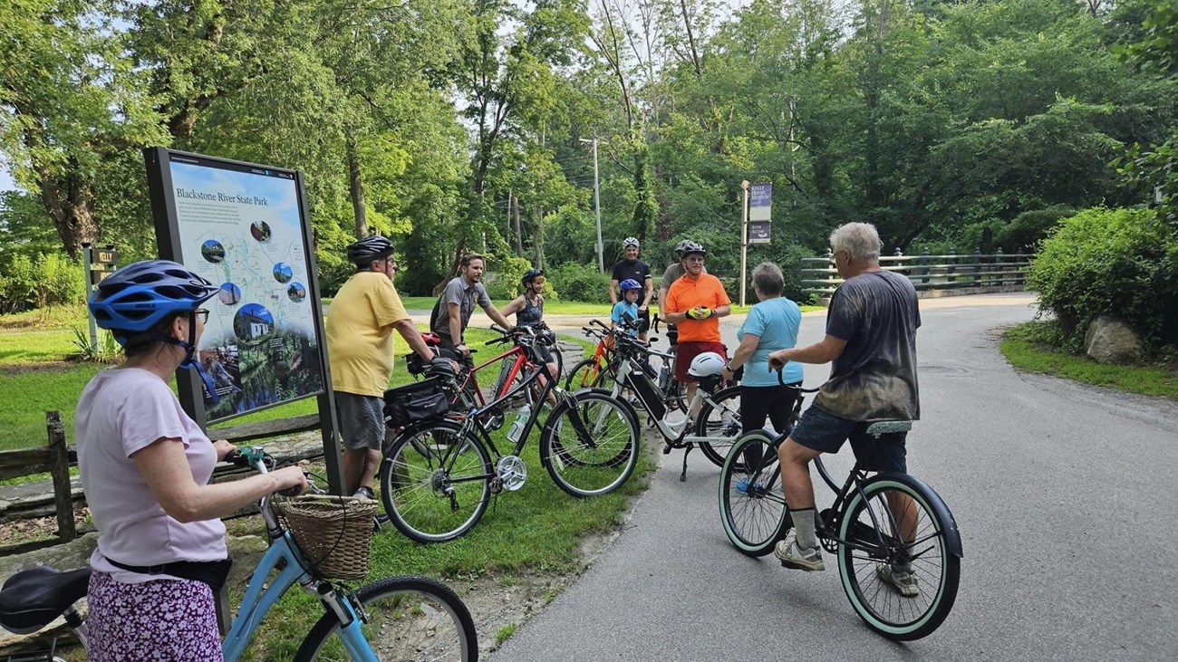 Group of cyclists gather wrong a Ranger and wayside panel for a guided bike ride