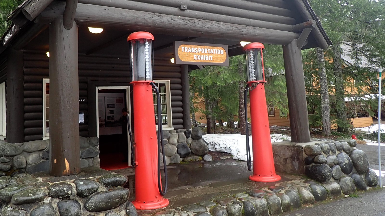 A small log building with two red historic gas pumps in front of it on a river rock island.