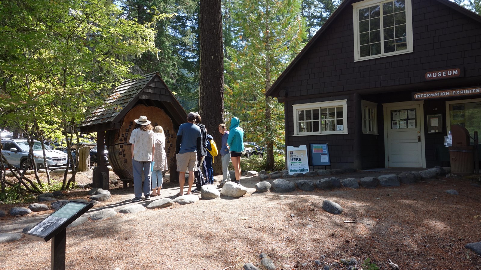 A small wayside panel in front of a wood cabin. Visitors stand on a path next to the cabin.