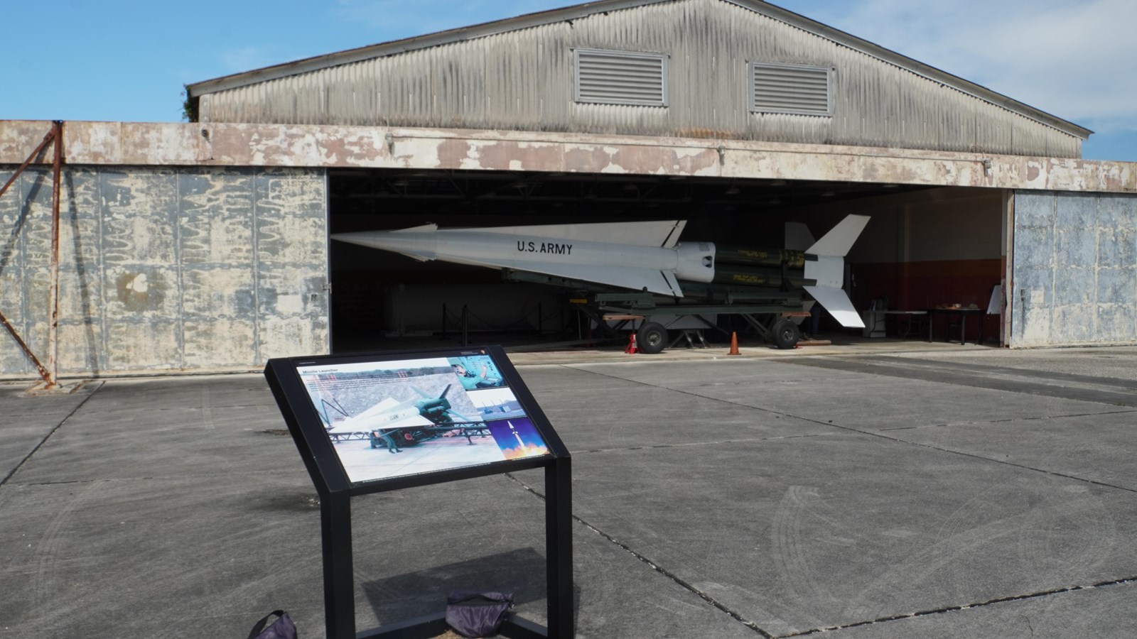 A colorful information wayside stands front of an open barn with a large green and white missile