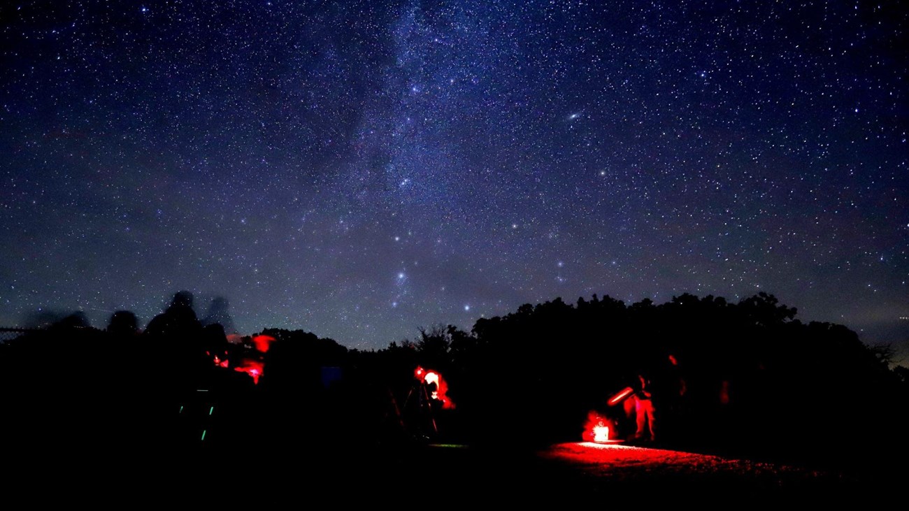 People standing around telescopes with red headlamps underneath a night sky