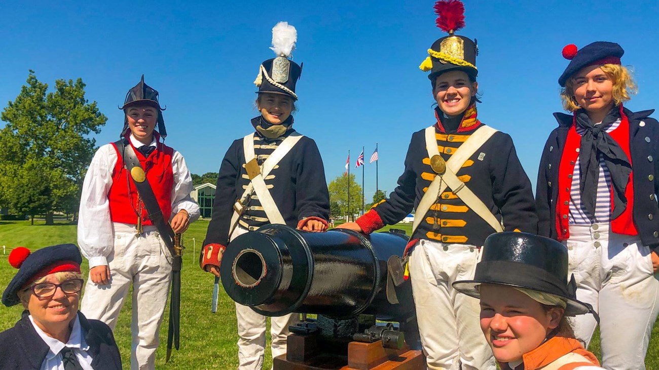 6 people dressed as soldier, marine, and sailors from the War of 1812 pose for a picture around a re