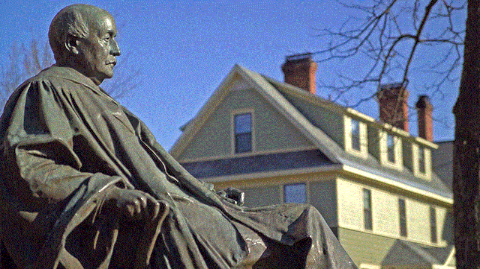 bronze statue of a male figure sitting on a chair sits atop a cement pedestal on a mowed lawn