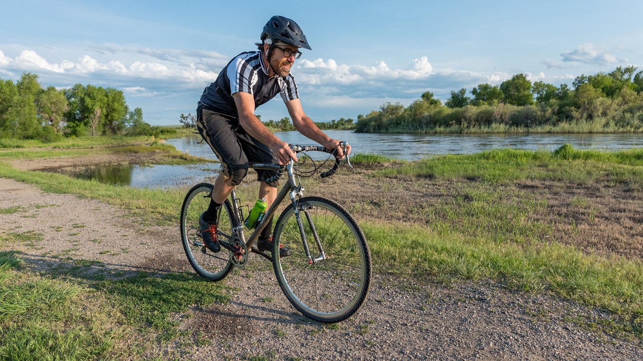 Cyclist biking dirt path along riverfront. Leafy trees and wetlands habitat in distance.