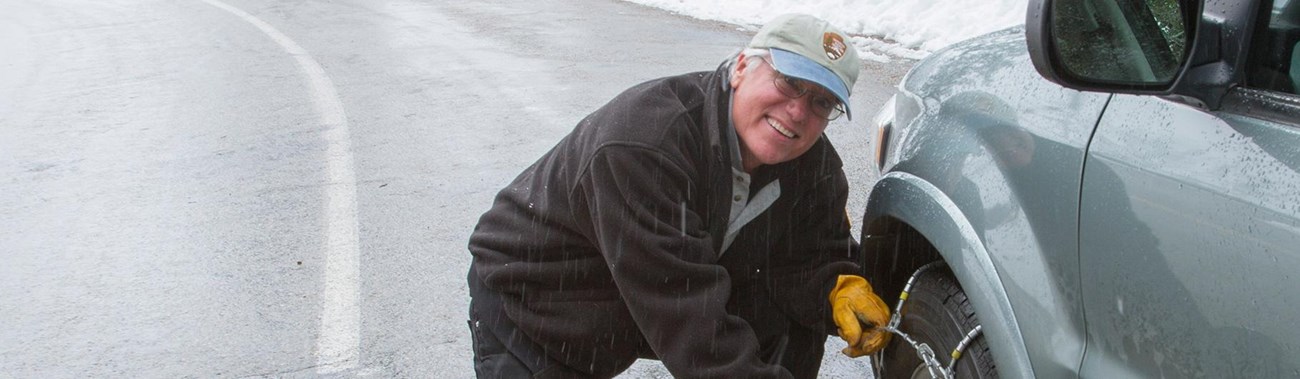 A man smiles at the camera as he finishes installing tire chains